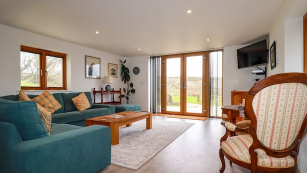 Ein Wohnzimmer mit Sofa, Couchtisch und Sessel. Ein Fernseher ist an der Wand montiert und es gibt ein Fenster mit Jalousien.