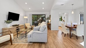 A modern living room with a sofa, coffee table, and TV. There is a dining area with a table and chairs. The room has wooden flooring and white walls. - Newly furnished Condo in Heart of Palm Desert! Pool, PickleBall! (Palm Desert)