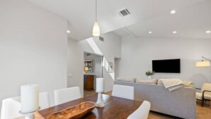 A modern living room with a grey sofa, a wooden dining table with white chairs, and a flat-screen TV mounted on the wall. - Newly furnished Condo in Heart of Palm Desert! Pool, PickleBall! (Palm Desert)
