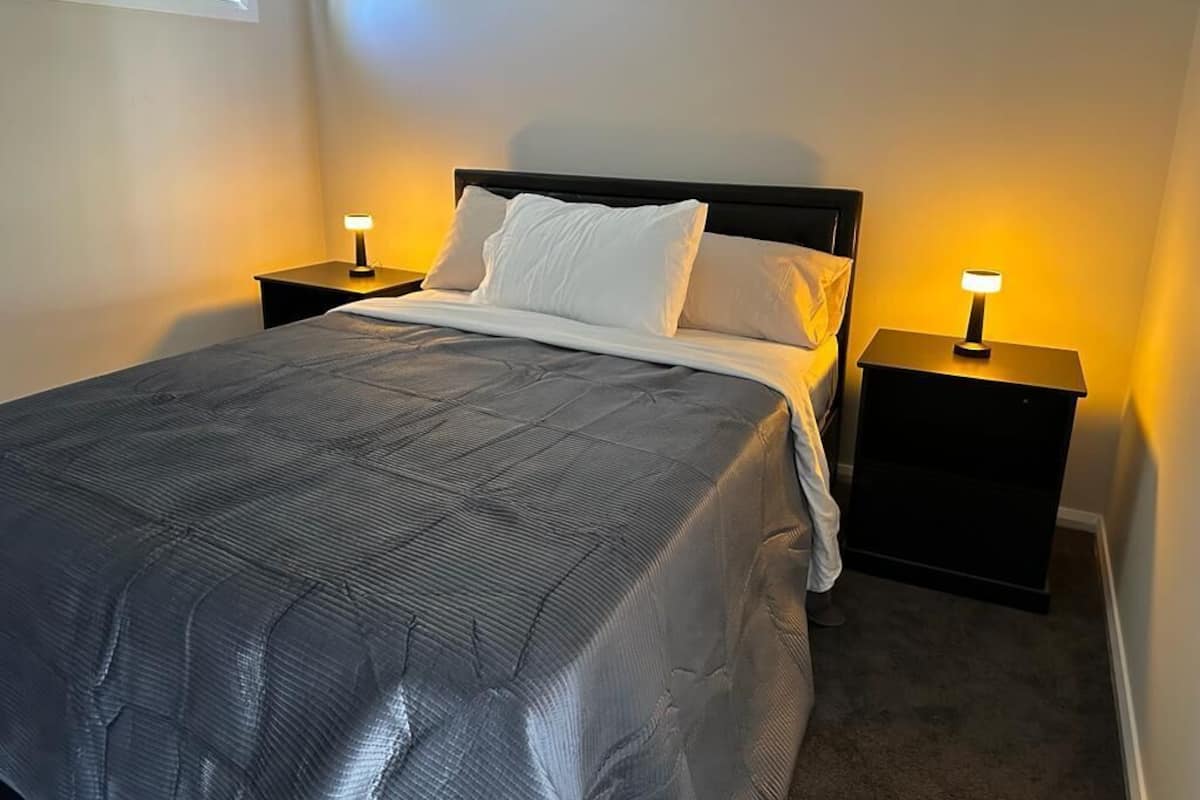 A neatly made bed with a dark gray comforter and white pillows, flanked by two bedside tables with lamps, in a room with a plain wall and carpeted floor.