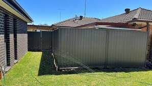 A fenced backyard with a lawn and a hose spraying water. - Granny Guest House Sydney Blue Mountain (Kingswood)