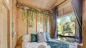 Une chambre dans une cabane en bois, avec un lit, des oreillers, une fenêtre avec des rideaux et des plantes grimpantes suspendues.