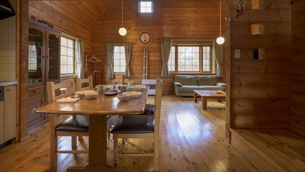 Interior de una cabaña de madera con una mesa de comedor, sillas, un sofá y una pequeña cocina.