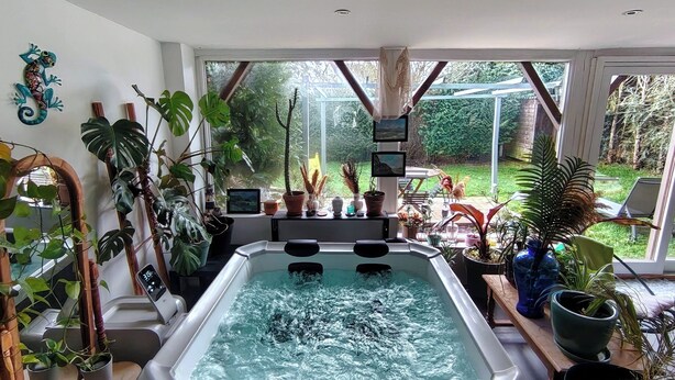 Ein Whirlpool in einem Raum mit großen Fenstern, umgeben von verschiedenen Kübelpflanzen und einem Laptop auf einem Tisch.