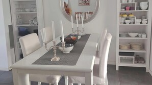Ein Essbereich mit einem weißen Tisch, gedeckt für sechs Personen, umgeben von weißen Stühlen. An der Wand hängt ein Spiegel, und rechts befindet sich ein weißes Regal mit Geschirr und Gläsern.