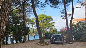 Ein Kiesweg führt zu einem geparkten Auto, umgeben von Bäumen und einem Gewässer im Hintergrund.