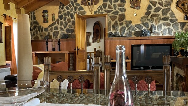 A rustic interior with a stone wall, wooden ceiling beams, a fireplace, a television, and a table with a glass vase. - Casa Obis - Casa Rural con Jardín, Barbacoa y Unas Espectaculares Vistas (Muñeca)