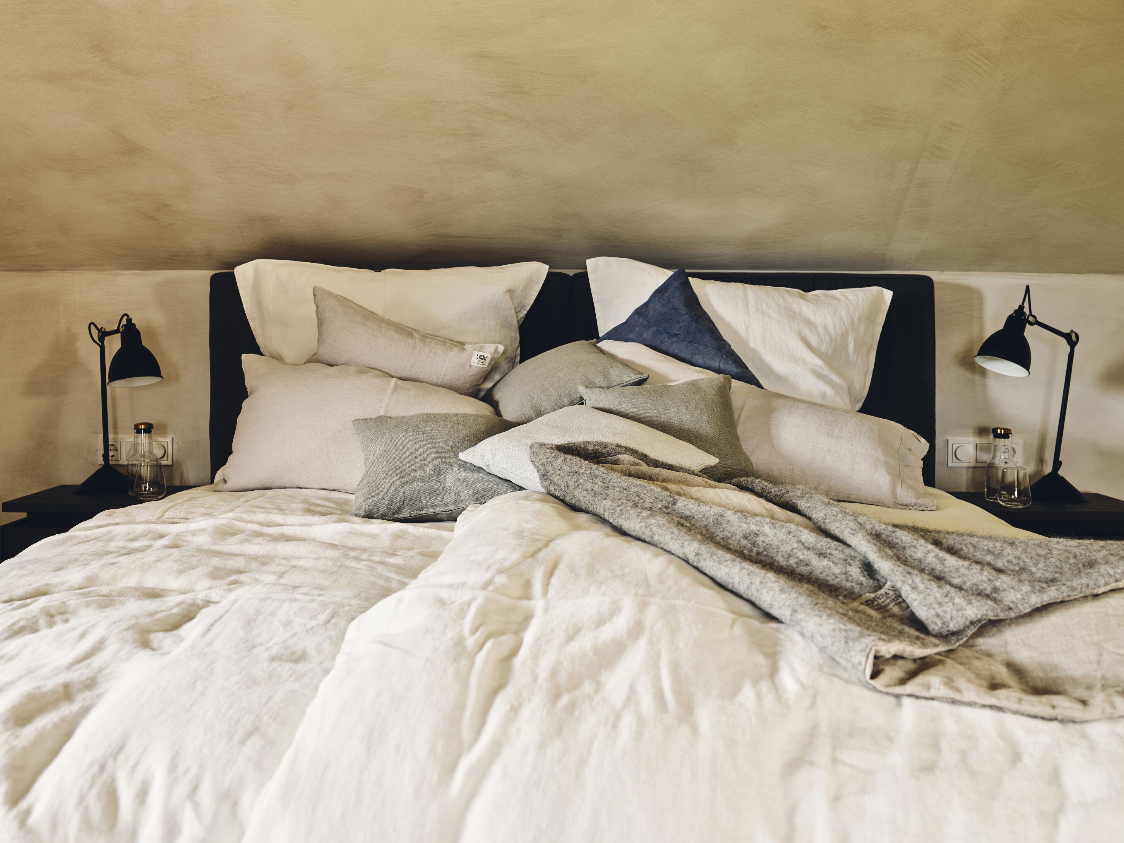 A bed with multiple pillows and a blanket, two bedside lamps, and a small bottle on a bedside table.
