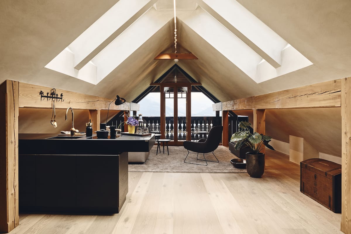 A spacious loft with a high ceiling, wooden beams, a black kitchen island, a dining area, and a view of the mountains.