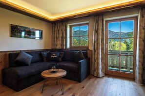 Une pièce chaleureuse avec un canapé aux couleurs sombres, une petite table en bois et une grande fenêtre donnant sur un paysage boisé.