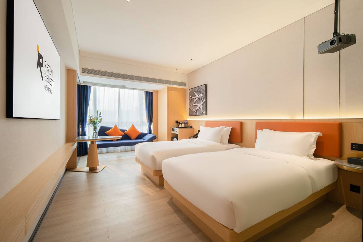 A hotel room with two beds, a desk, a chair, a sofa, and a large window with curtains.