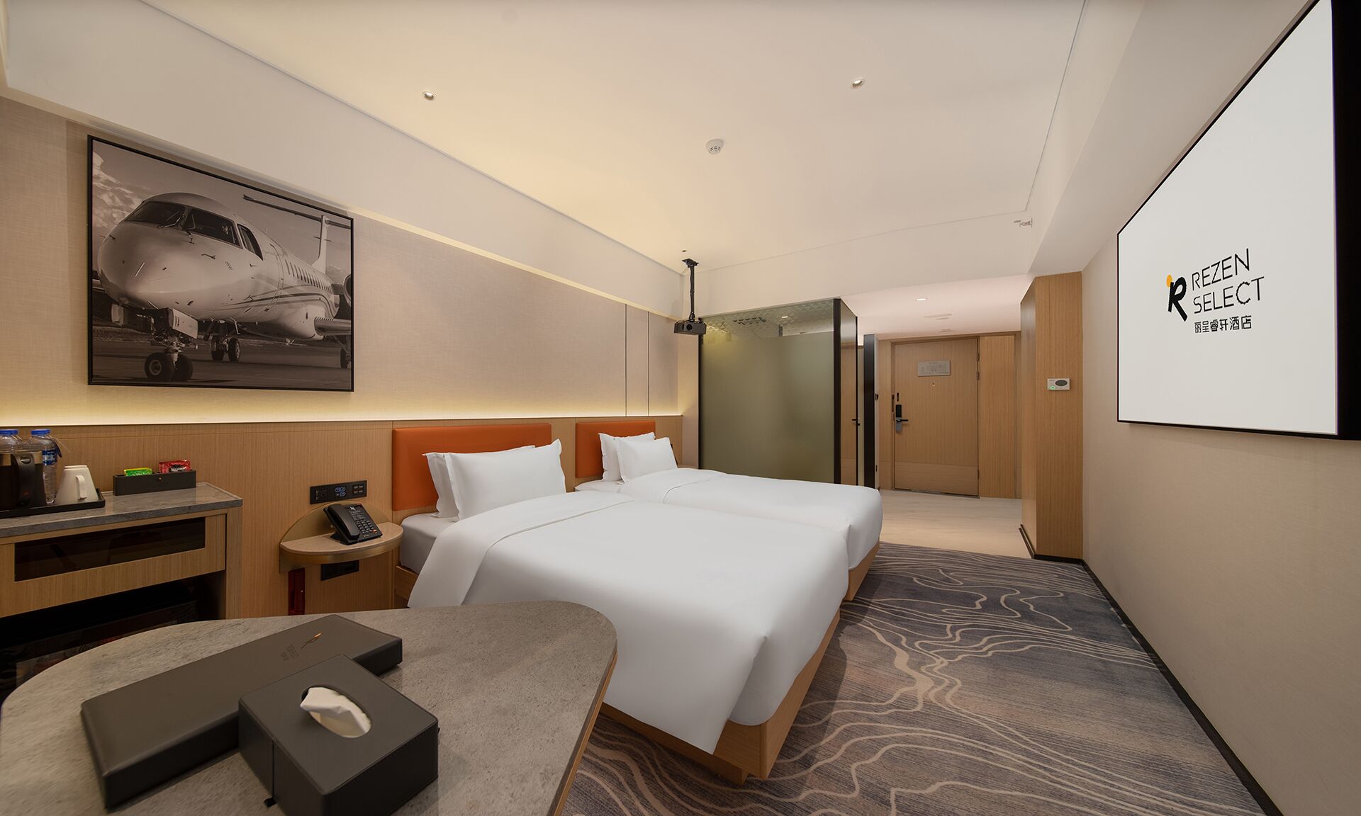 Habitación de hotel con una cama grande, un escritorio con una computadora, un teléfono y una pequeña mesa con una caja de pañuelos. En la pared, hay un cuadro de un avión y un cartel con la marca del hotel.