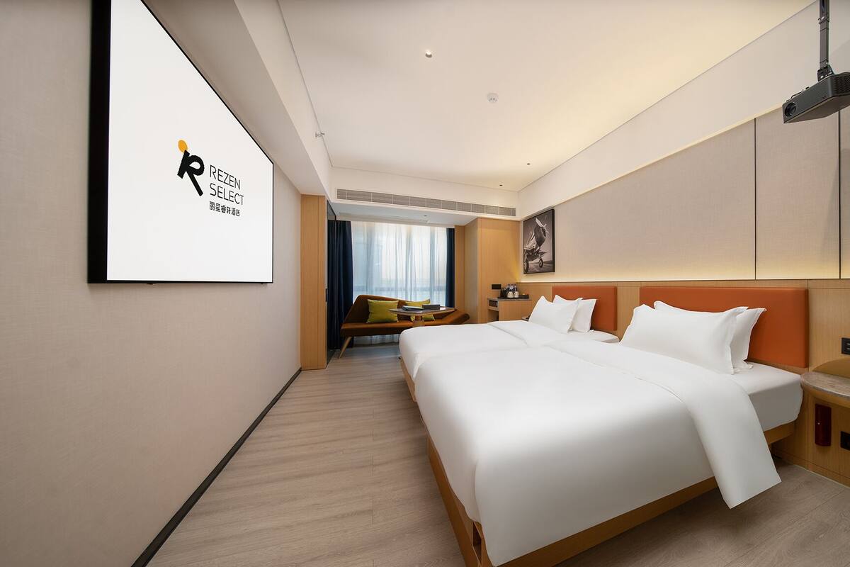 A modern hotel room with a large bed, a seating area with a sofa and armchairs, a desk, and a large window with curtains.