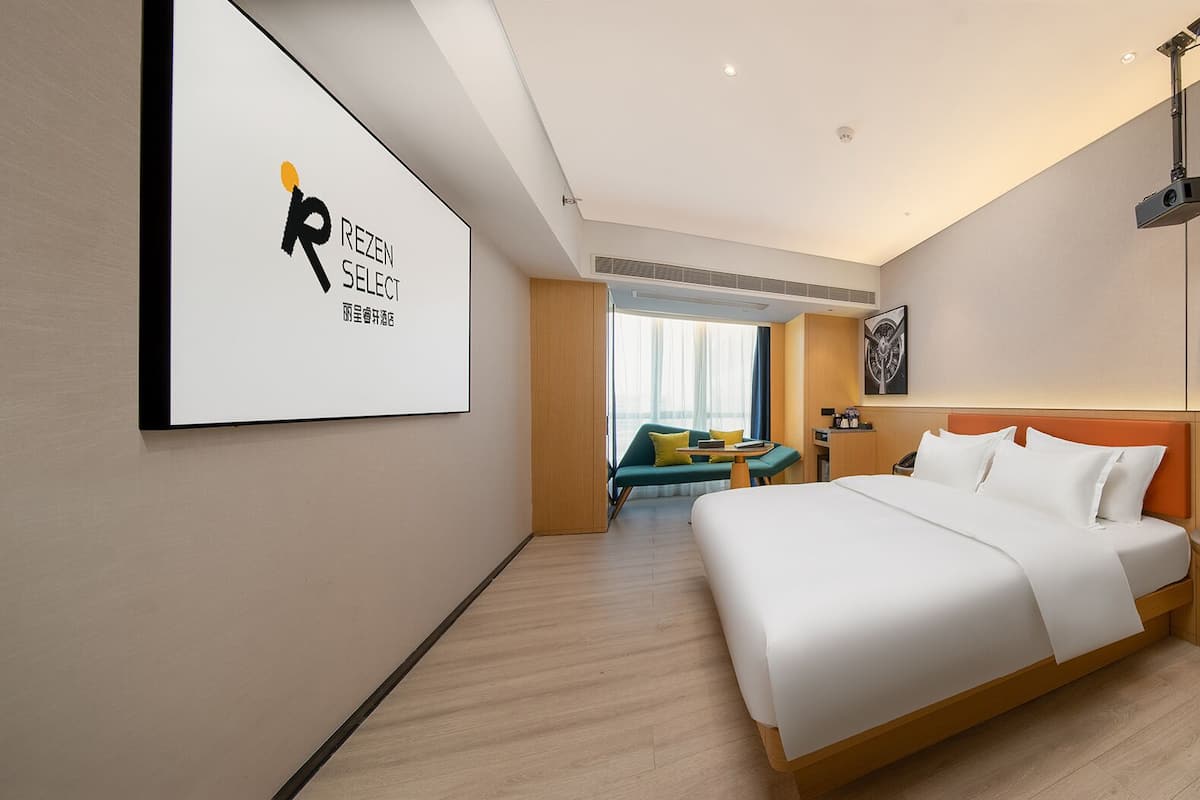 A modern hotel room with a large bed, a seating area with a sofa and table, and a wall-mounted projector.
