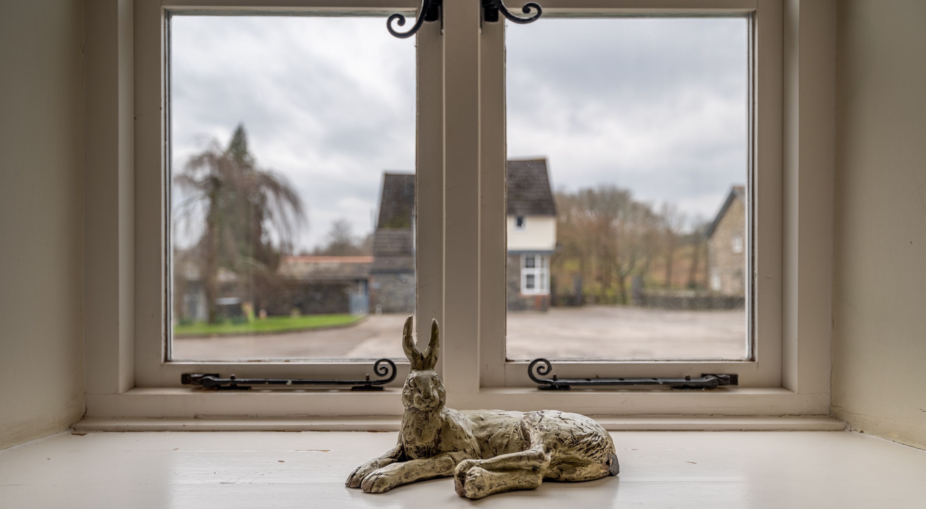 A window with a sculpture of a reclining animal on the sill, overlooking a residential street with houses and trees.