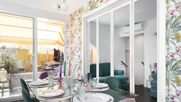 Un coin repas avec une table en verre dressée pour quatre personnes, entourée de chaises de couleur turquoise. Un grand miroir reflète la pièce, et un papier peint à motifs floraux orne les murs.