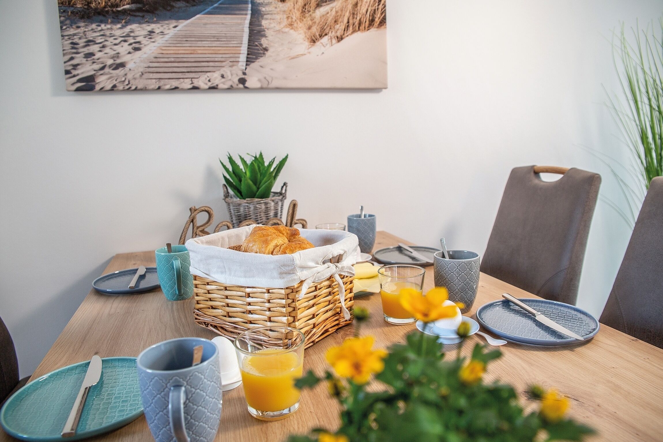 A breakfast table with a wicker basket, a plant, a croissant, a glass of orange juice, and a plate with a knife and fork.
