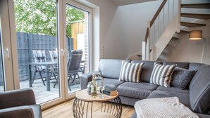 A modern living room with a grey sofa, striped pillows, a round coffee table, and a view of an outdoor patio with a table and chairs. - Deichkajüte 2 - Diese Neuwertige Doppelhaushälfte Bietet Ihnen Höchsten Komfort (Carolinensiel)