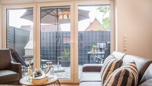 A modern living room with a sofa, armchairs, and a coffee table. There is a sliding glass door leading to a patio with outdoor furniture. - Deichkajüte 2 - Diese Neuwertige Doppelhaushälfte Bietet Ihnen Höchsten Komfort (Carolinensiel)