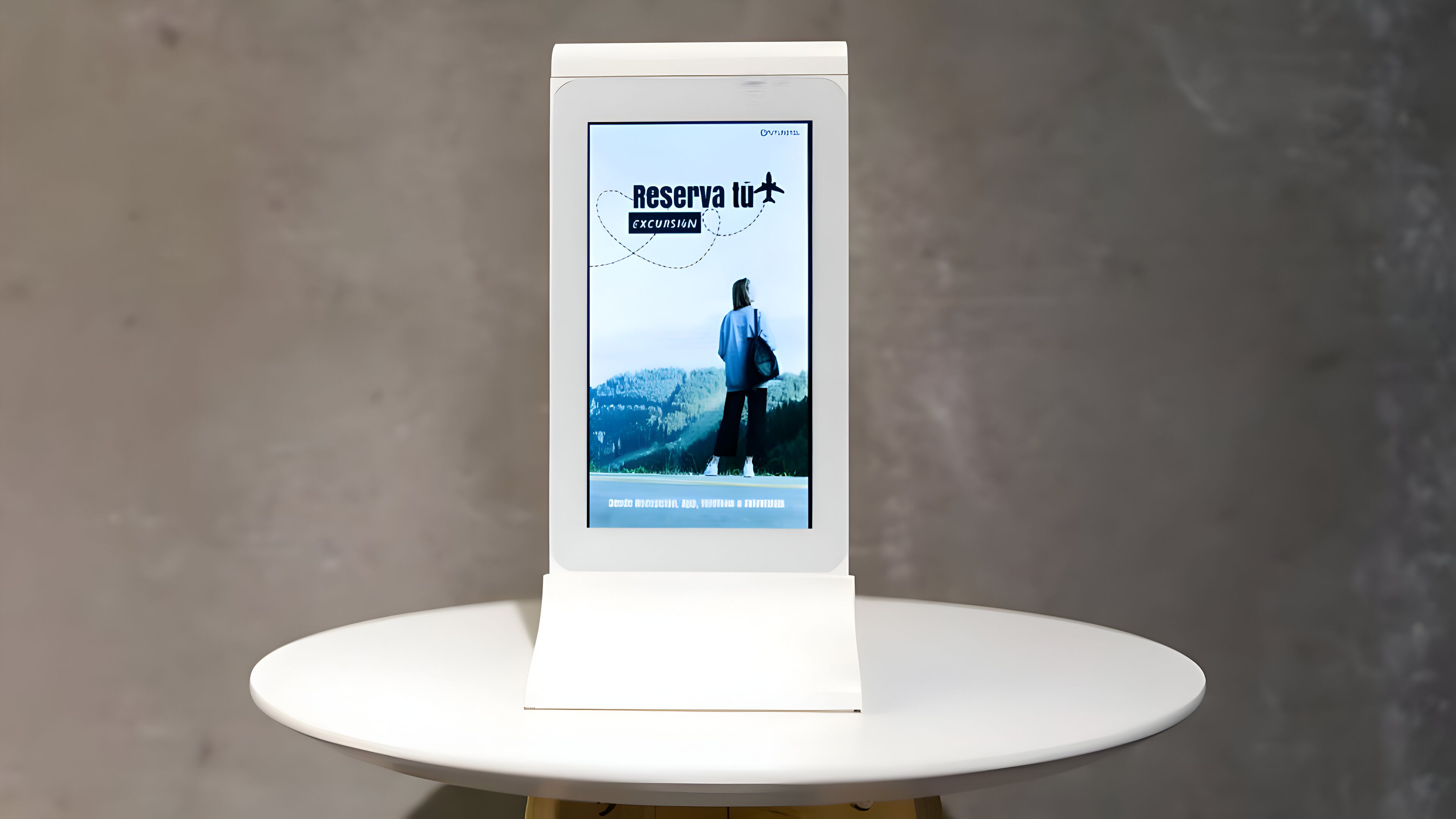 Una pantalla digital sobre una mesa blanca que muestra un anuncio de viajes con una persona caminando por un camino con montañas al fondo.