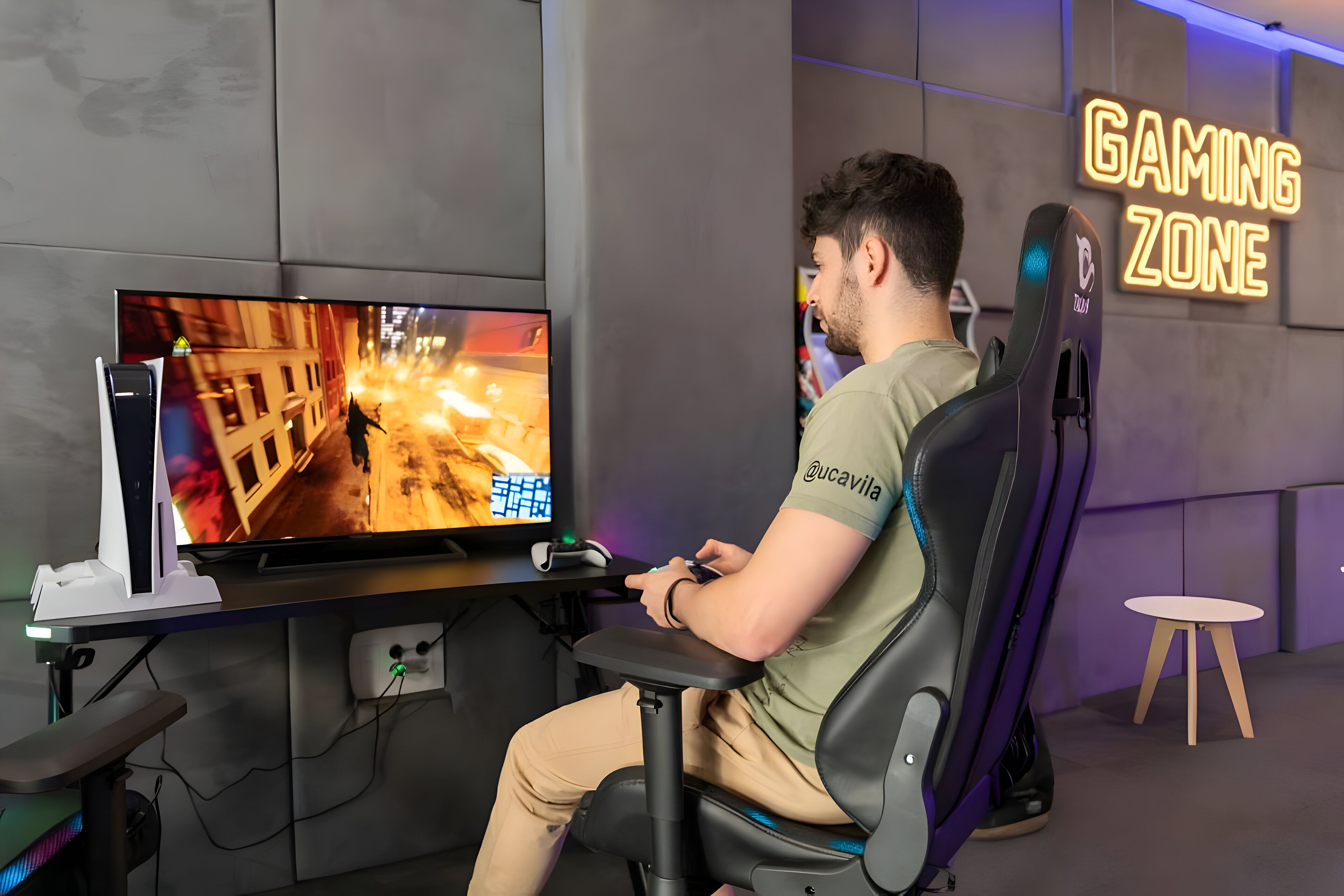 A person seated in a gaming chair at a desk with a monitor displaying a video game. A neon sign reads "GAMING ZONE."