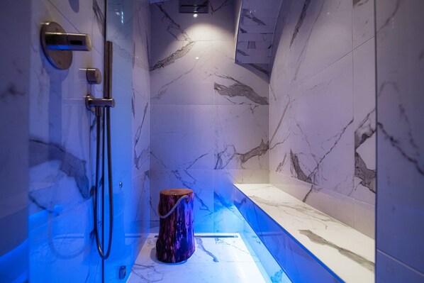 Un baño moderno con paredes de mármol, ducha integrada y bañera iluminada con luz azul.