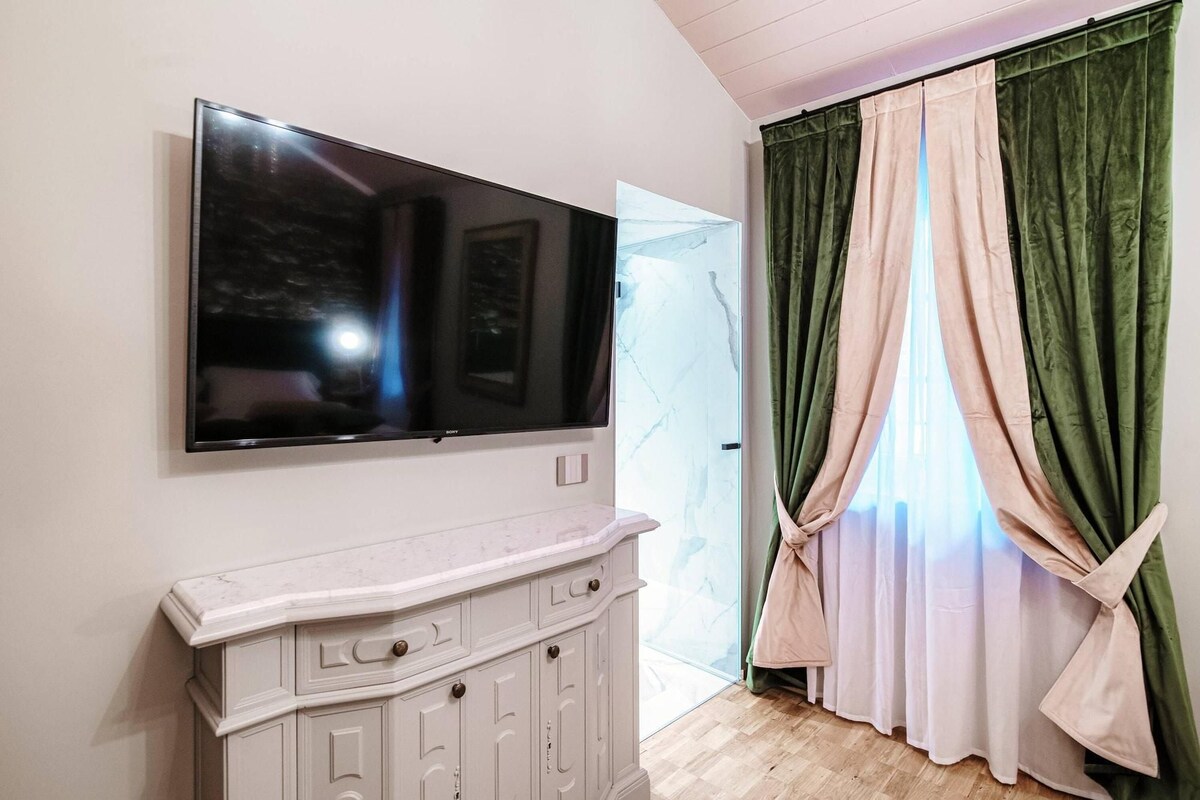 A room with a flat-screen TV mounted on the wall, a white cabinet below it, and a window with green and white curtains.