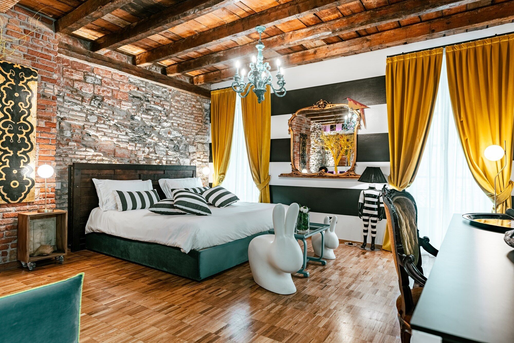 Une chambre avec un mur en briques, des poutres en bois au plafond, un lit avec des coussins à rayures, un miroir et des rideaux jaunes.