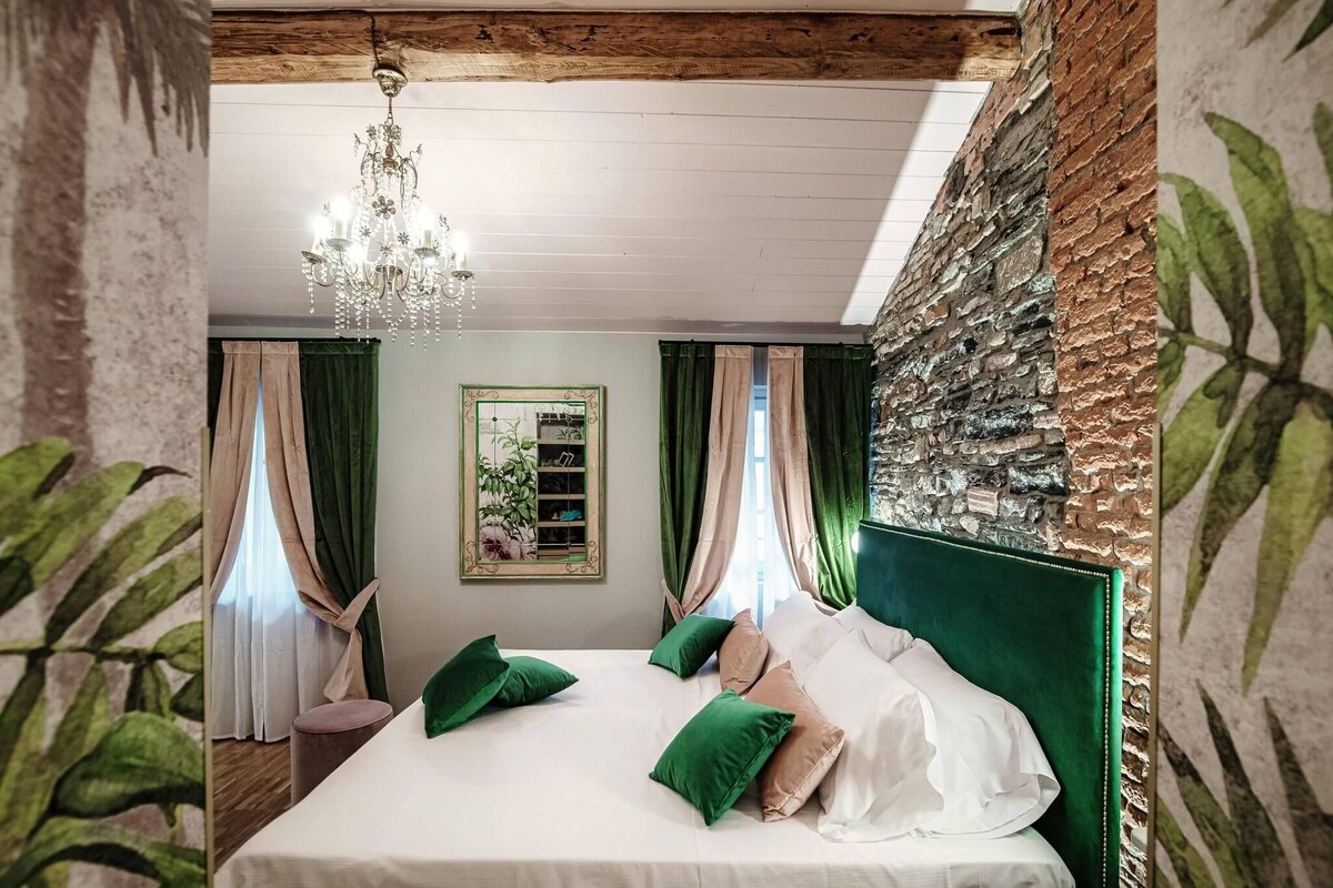 A bedroom with a large bed, green and beige pillows, a chandelier, a mirror, and a green headboard.