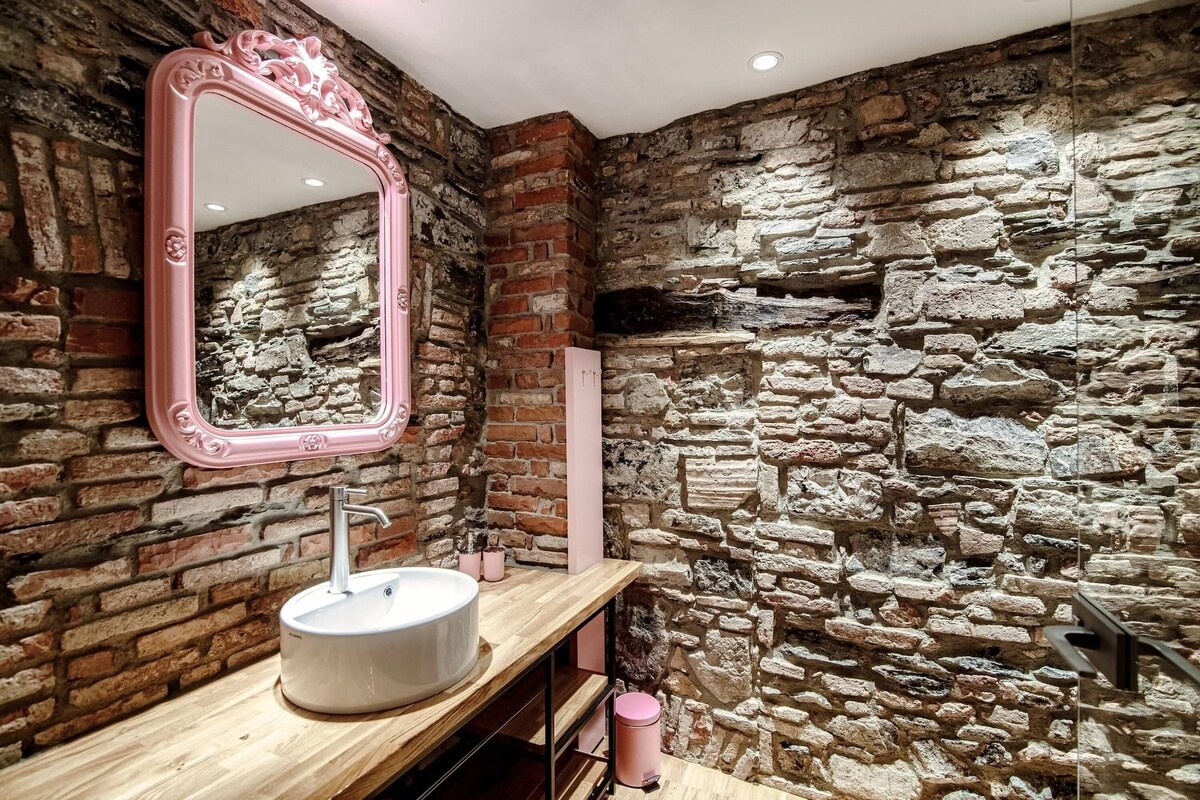 A bathroom with a stone wall, a pink-framed mirror, a wooden vanity, and a modern sink.