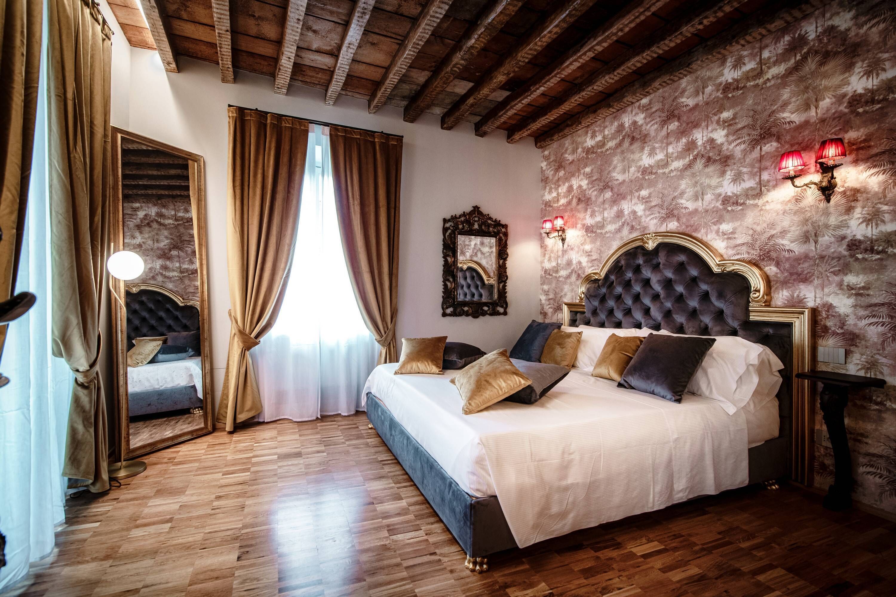A bedroom with a large bed, wooden flooring, a mirror, and a wall with a floral pattern.