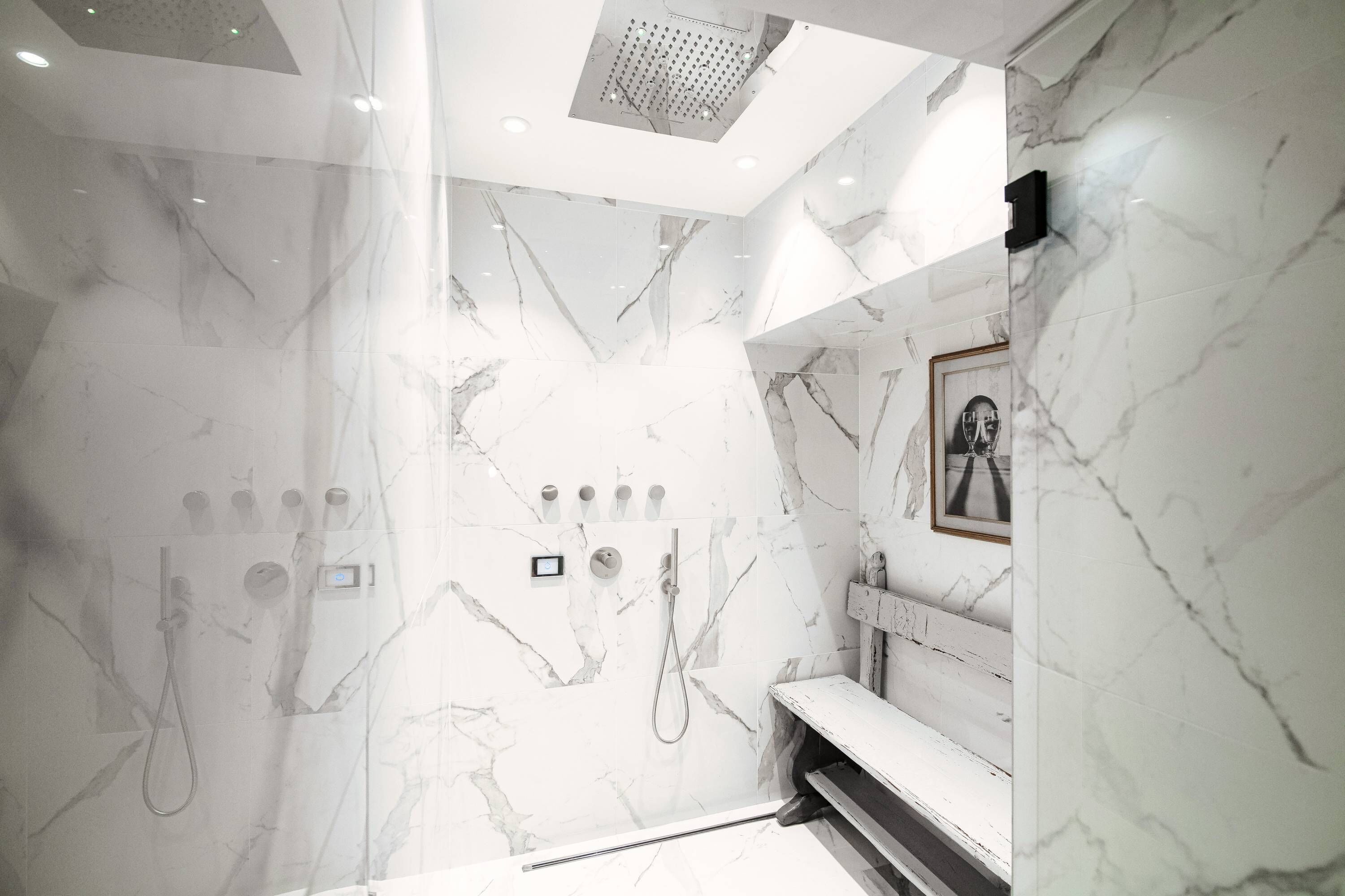 Un baño moderno con pared de ducha de mármol, un banco y un cuadro enmarcado.