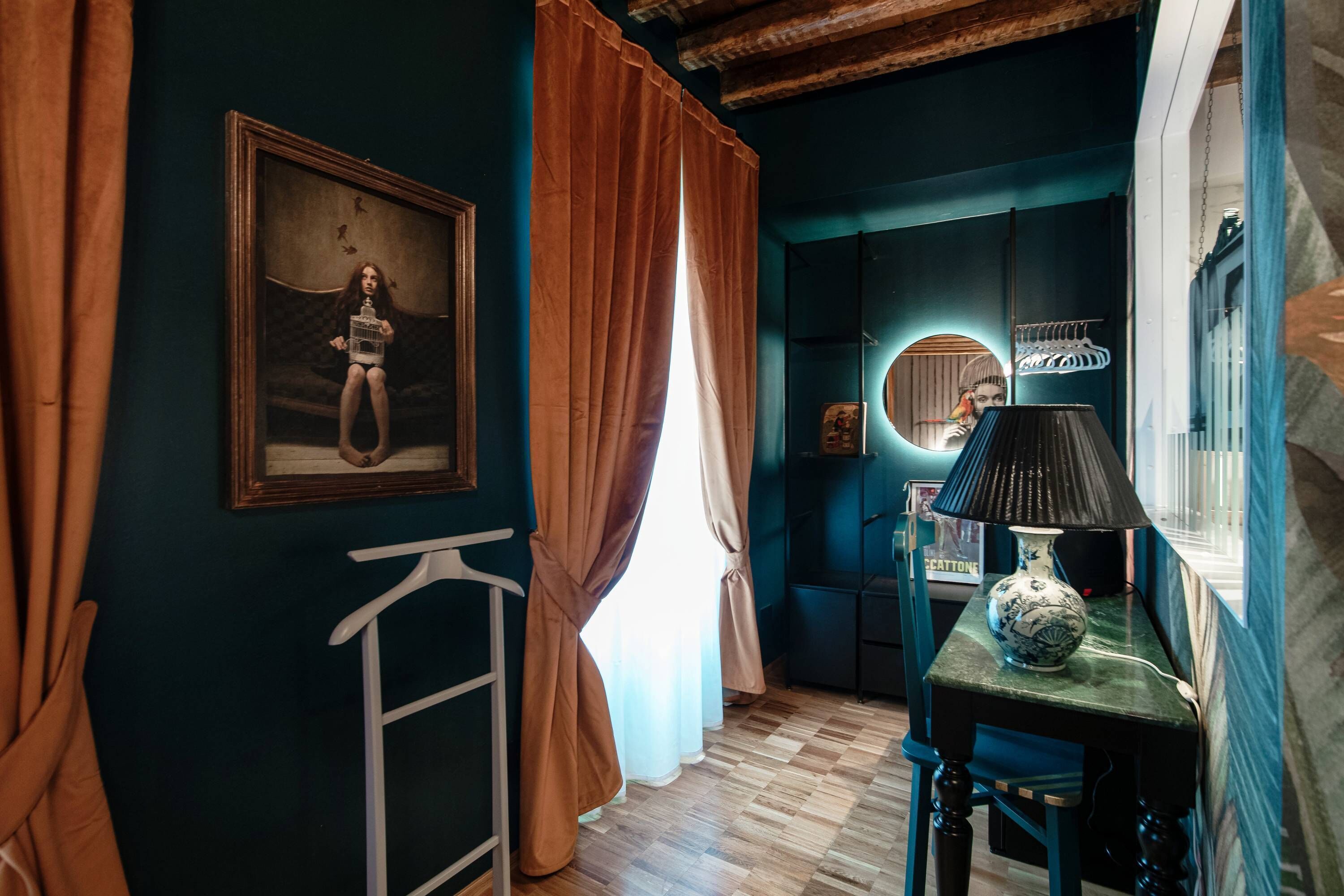A room with a wooden floor, a table with a lamp, a framed picture on the wall, and a window with curtains.