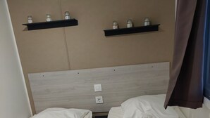 Ein Zimmer mit zwei Betten, beide mit weißer Bettwäsche und auf einem Bett eine violette Decke. Darüber befinden sich zwei Regale mit Gläsern darauf.