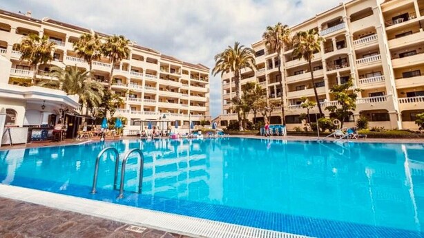 Een hotel met een groot zwembad, omringd door palmbomen en meerdere balkons.