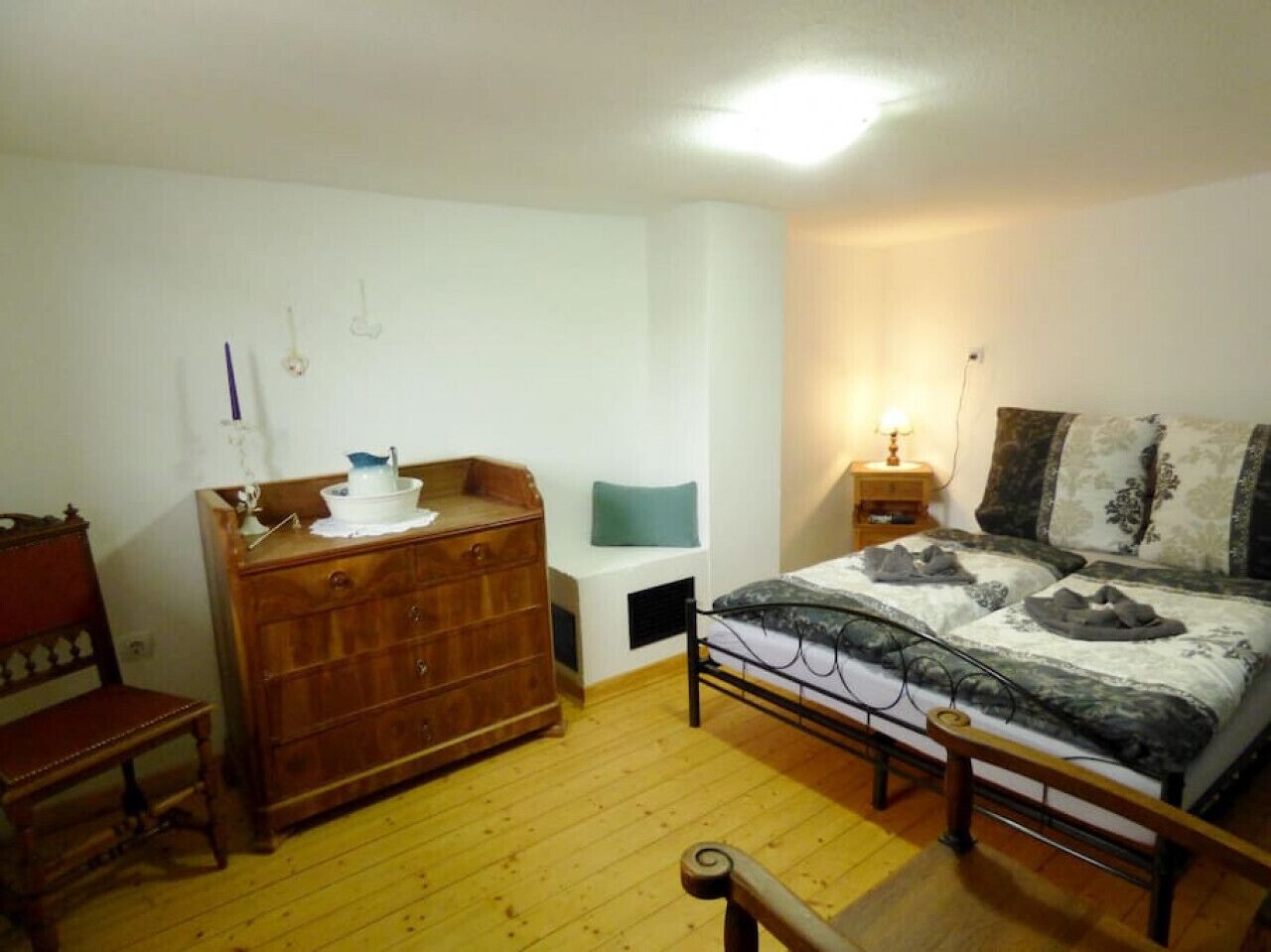 A bedroom with a wooden dresser, a bed with patterned bedding, a bedside table with a lamp, and a chair.