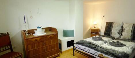 A bedroom with a wooden dresser, a bed with patterned bedding, a bedside table with a lamp, and a chair.