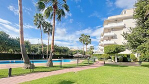 A pool area with palm trees, a brick pathway, and a building with balconies. - Global Vacacional: Practical flat in Canet Beach (Canet d´en berenguer)