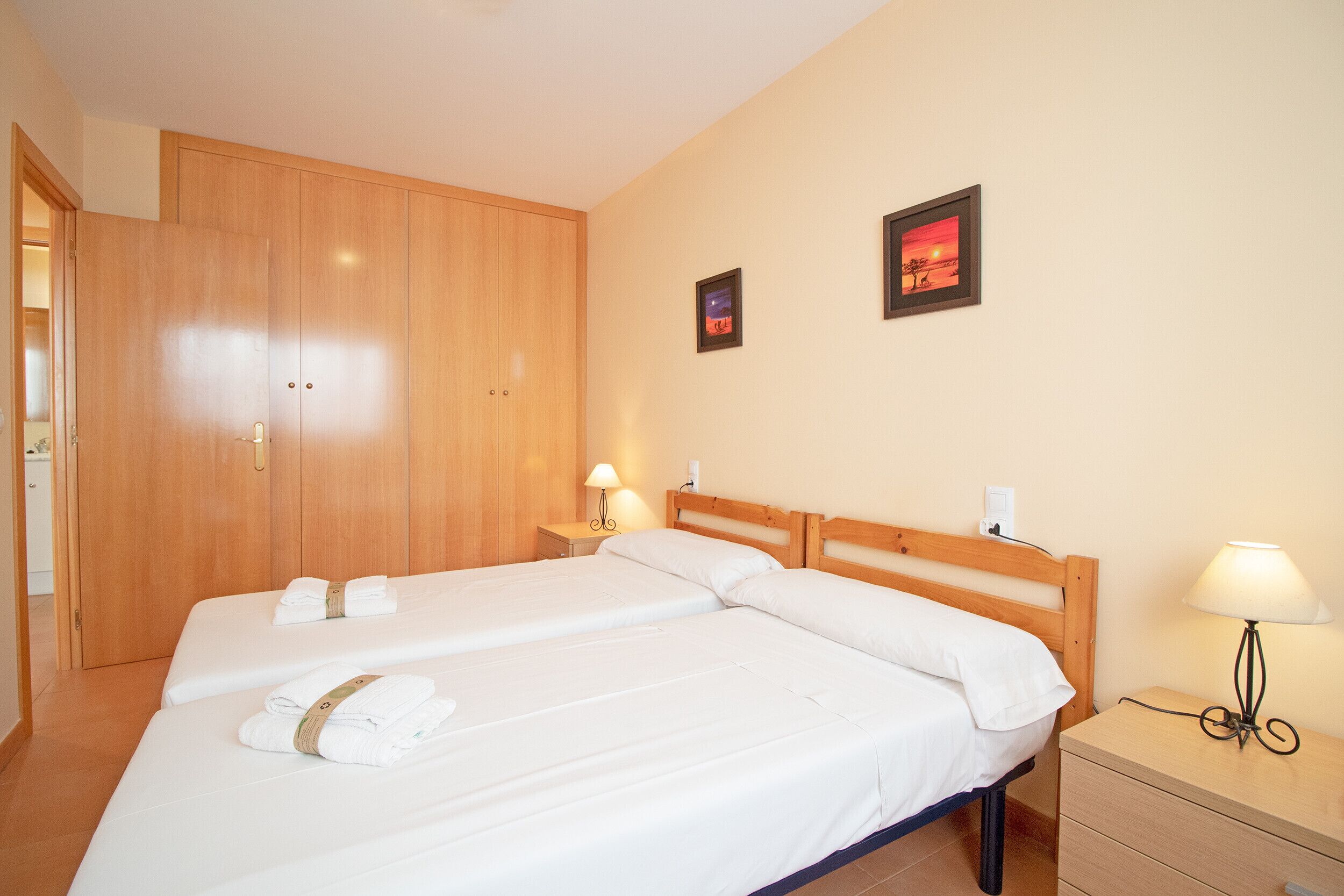 Habitación de hotel con dos camas, un armario de madera, una mesita de noche con lámpara y dos cuadros enmarcados en la pared.