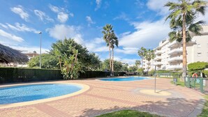 A swimming pool area with a brick pathway, palm trees, and a building in the background. - Global Vacacional: Practical flat in Canet Beach (Canet d´en berenguer)