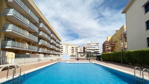 Una piscina rodeada de edificios de varios pisos con balcones, cielo despejado y un seto a un lado.