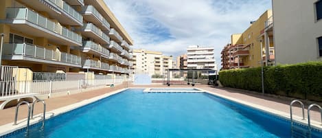 Una piscina rodeada de edificios de varios pisos con balcones, cielo despejado y un seto a un lado.