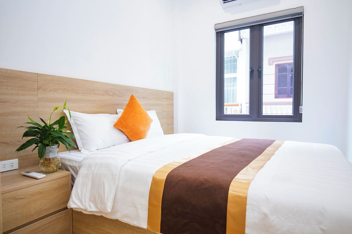 Una cama bien hecha con cabecera de madera, una planta en la mesita de noche y una ventana con vistas a edificios.