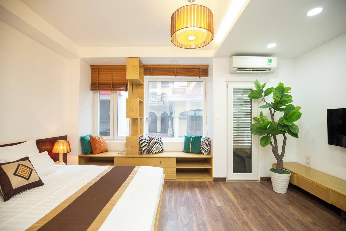 Habitación de hotel con cama, mesita de noche, lámpara, ventana con persianas, banco integrado, planta en maceta y televisor de pantalla plana.