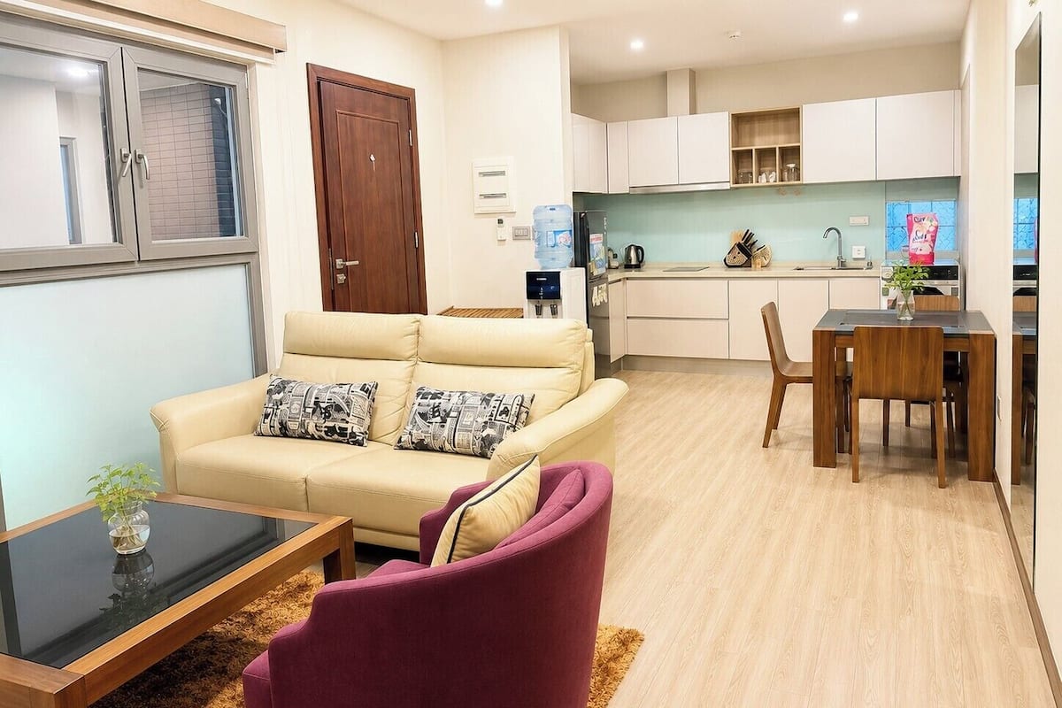 Una sala de estar con un sofá, una mesa de centro y una silla. Hay una zona de cocina con armarios y una mesa de comedor.