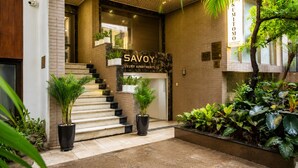 A modern building entrance with a staircase leading to a lobby area, potted plants, and a sign for 'SAVOY LUXURY APARTMENTS.'