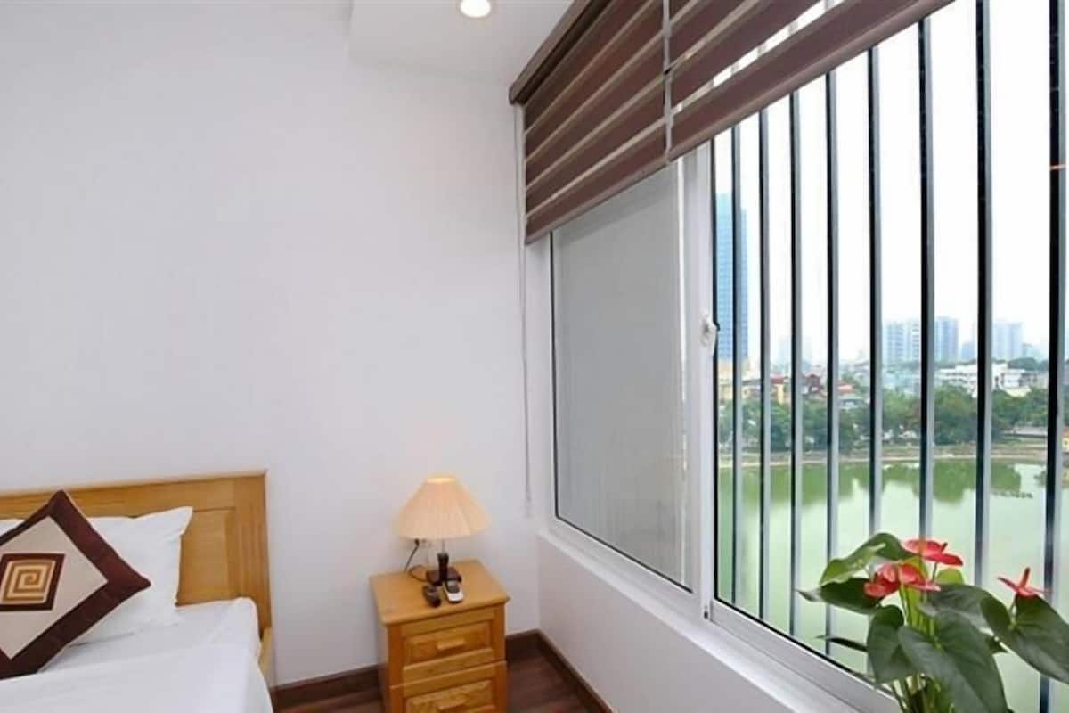 Habitación de hotel con cama, mesita de noche con lámpara y un amplio ventanal con vista a la ciudad.