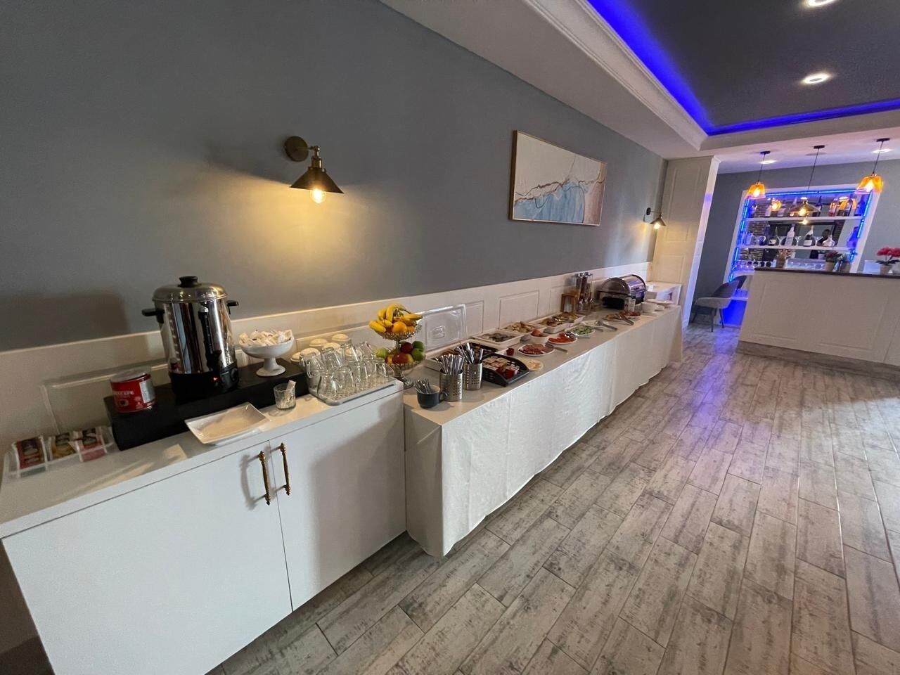 A buffet setup with a variety of food items and utensils, a refrigerator, and a bar area with blue lighting.