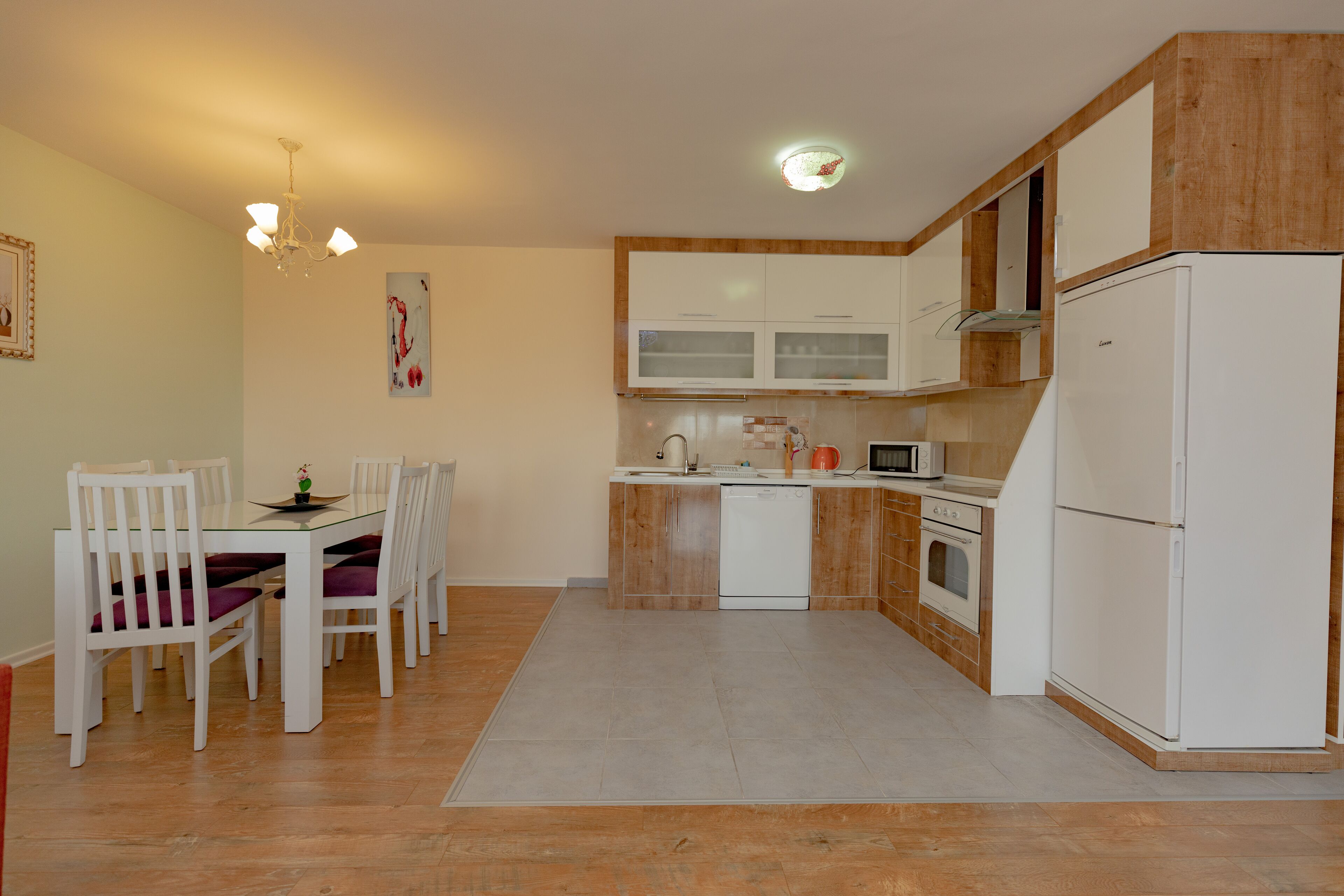 A modern kitchen with white cabinets, a dishwasher, a microwave, and a refrigerator. There is a dining table with chairs and a chandelier.