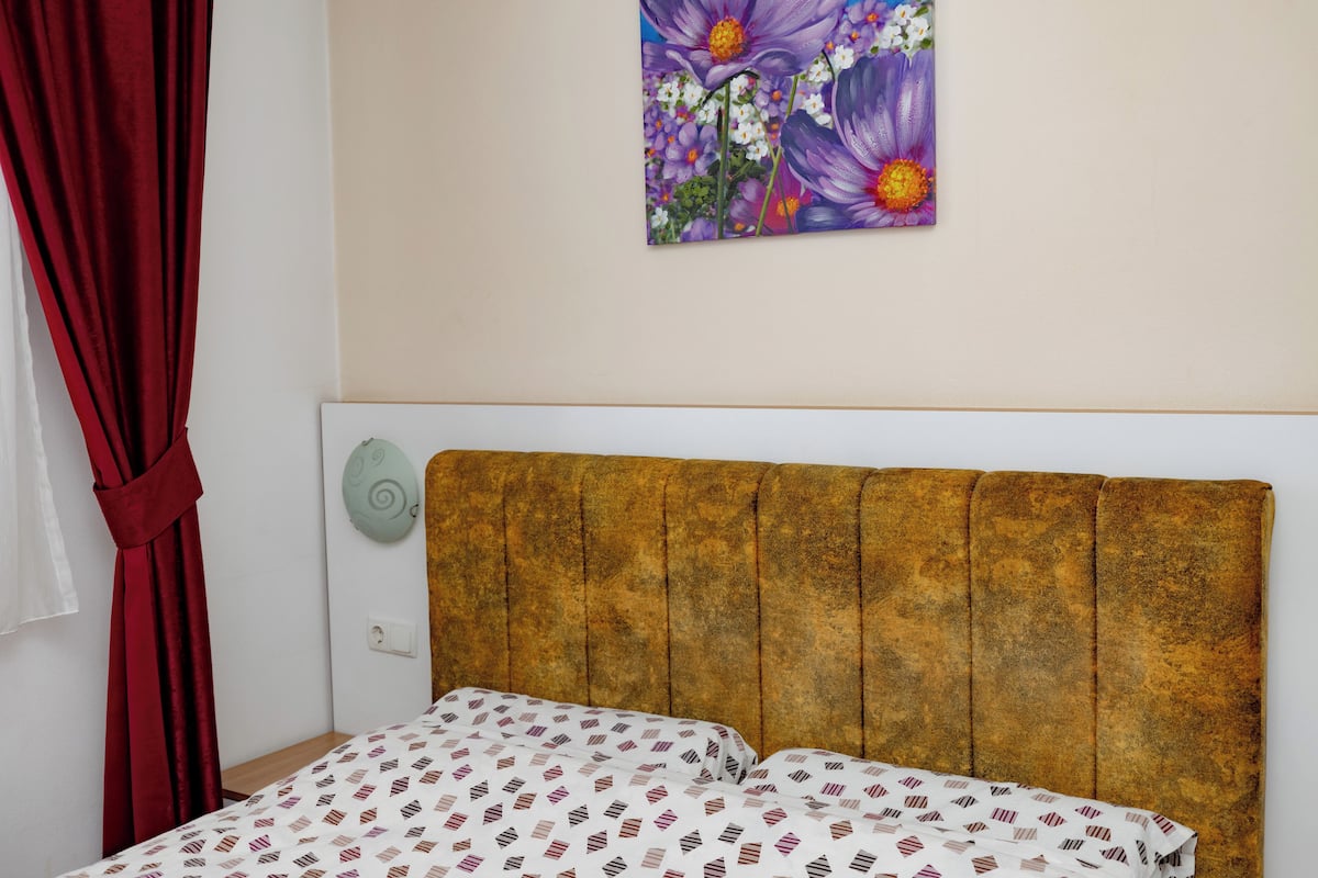 Ein Schlafzimmer mit einem Kopfteil, einem Bett mit gemusterten Betttextilien, einem roten Vorhang und einem floralen Gemälde an der Wand.
